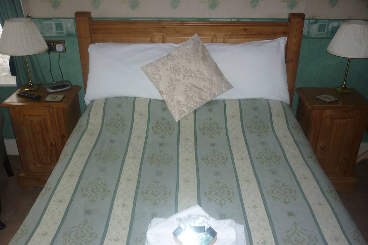 Una cama bien hecha con una colcha estampada, una almohada decorativa y una toalla doblada encima. Hay dos mesitas de noche, con lámparas y un control remoto sobre una de ellas.