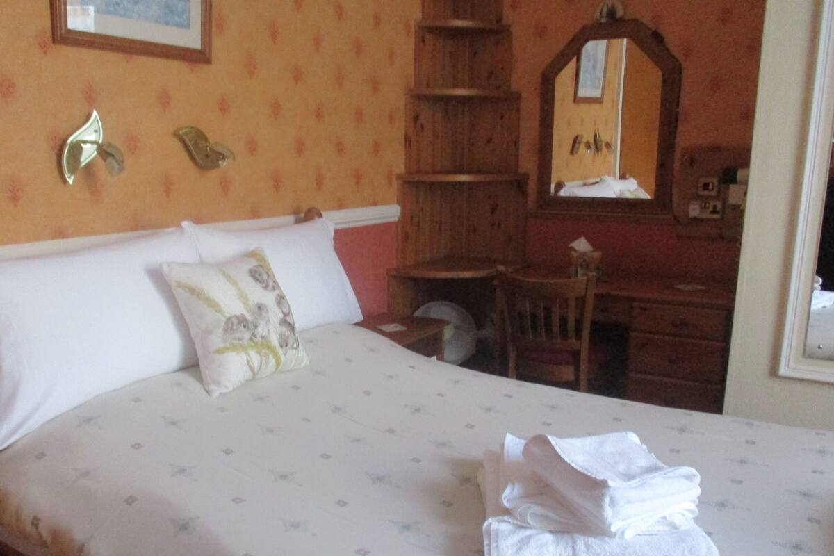 Una cama bien tendida con una almohada y una toalla doblada, un aparador de madera y un espejo.