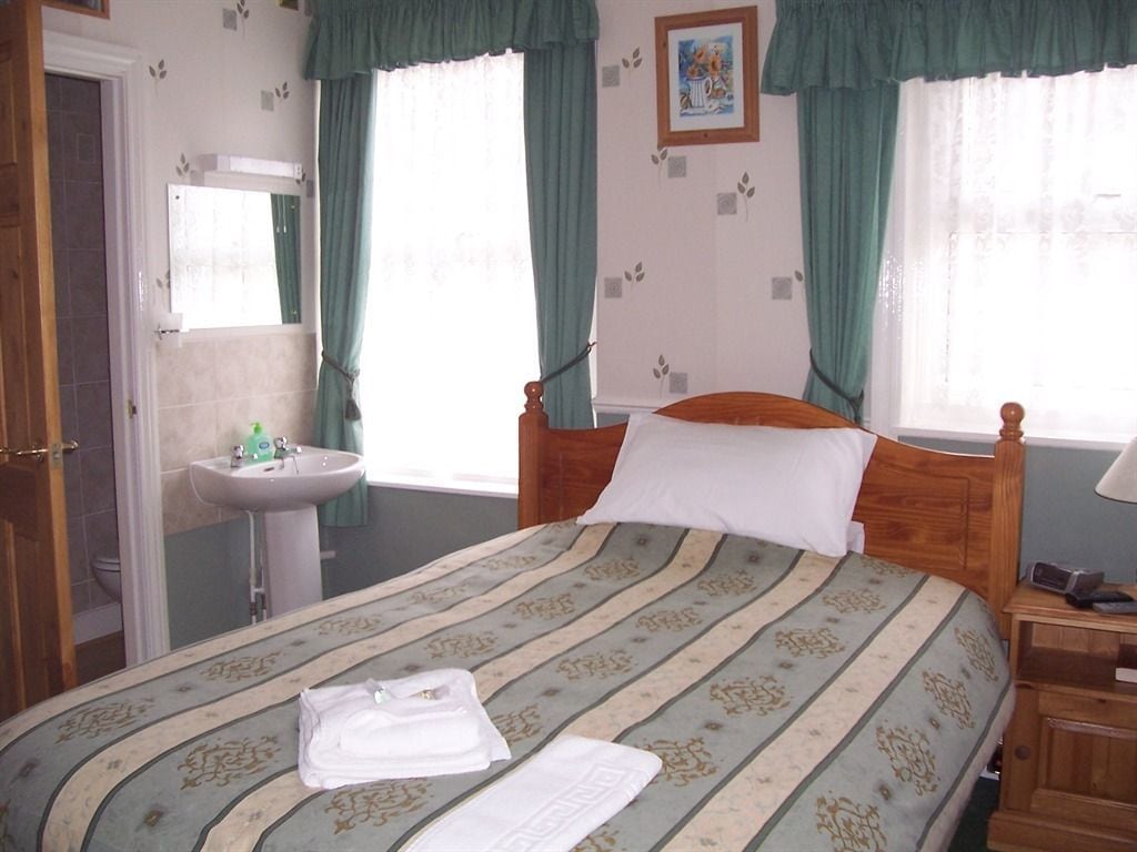 Un dormitorio con cama, mesita de noche, cortinas en la ventana, un cuadro en la pared y un baño con lavabo y espejo.