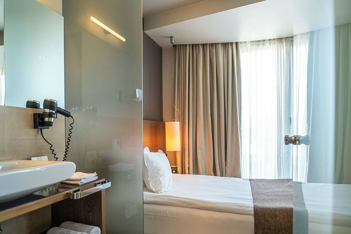 A hotel room with a glass partition, a bed, a bedside table with a lamp, a bathroom with a sink and a hairdryer, and a window with curtains.