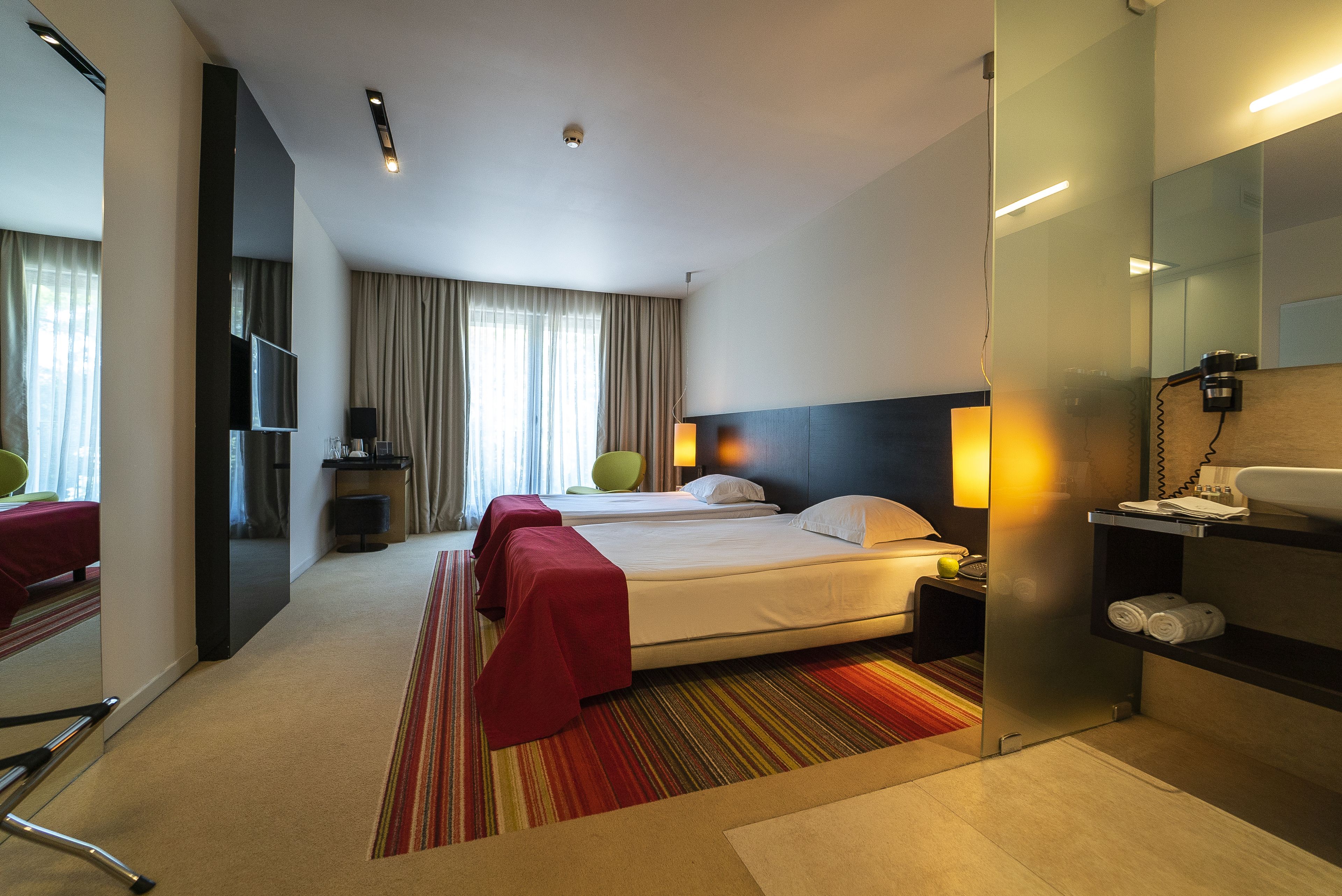 Ett hotellrum med två sängar, en TV, ett skrivbord med en lampa, en spegel och ett badrum med handfat och handduksställ.