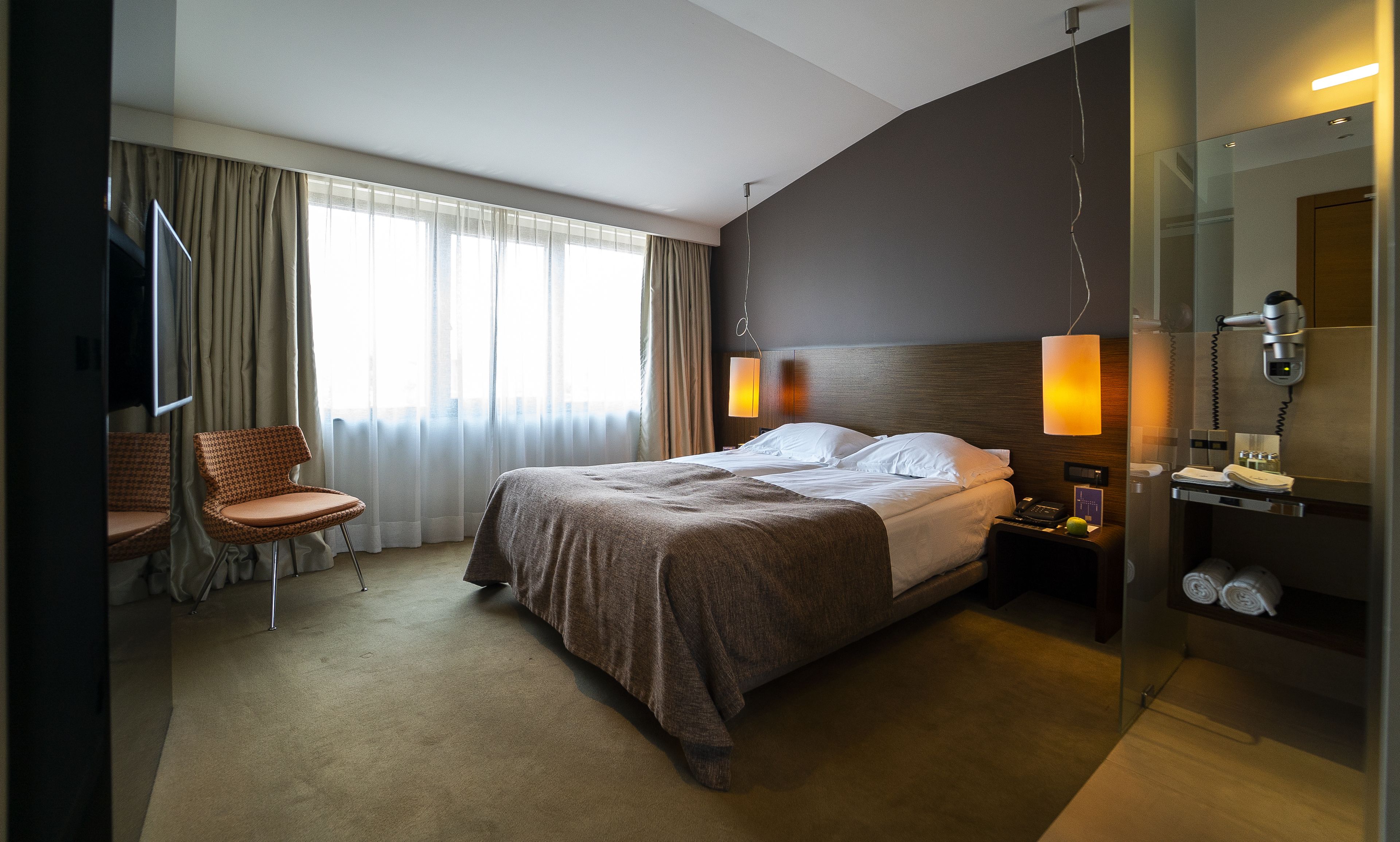 Habitación de hotel con cama, una silla, mesita de noche con lámpara, espejo y zona de ducha.