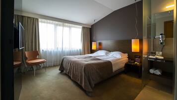 Habitación de hotel con cama, una silla, mesita de noche con lámpara, espejo y zona de ducha.