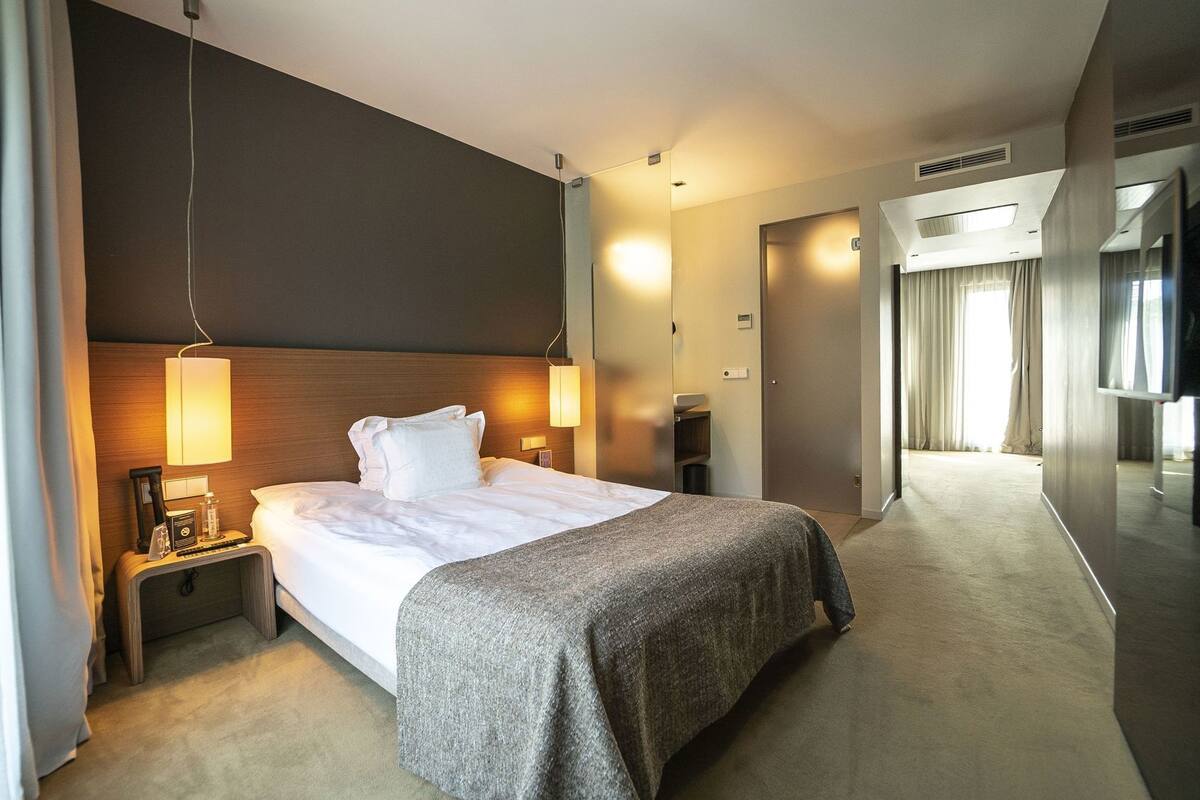 A hotel room with a bed, bedside lamps, a nightstand, a carpeted floor, and a hallway leading to a bathroom.