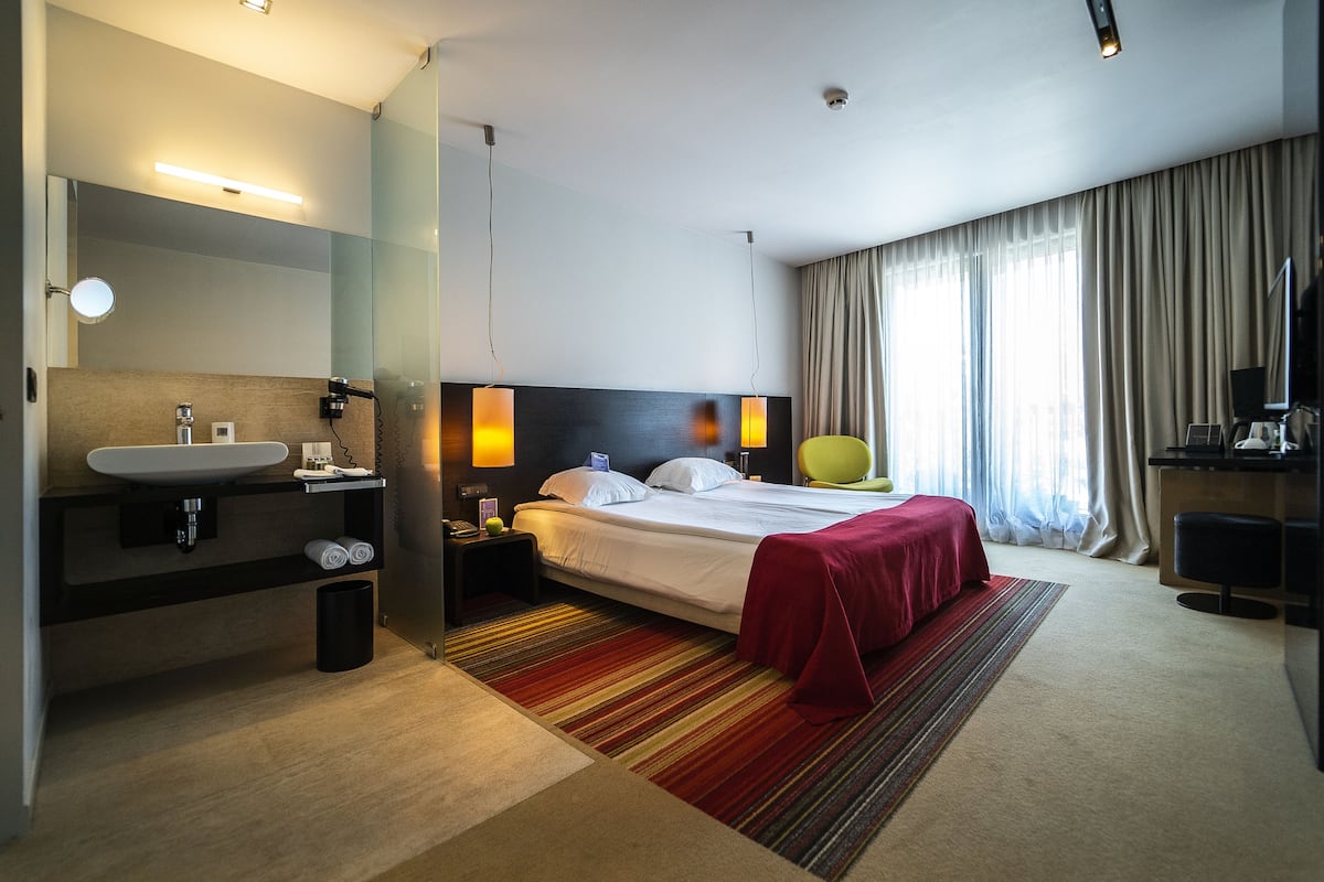 A modern hotel room with a large bed, a bedside table with a lamp, a bathroom with a sink and mirror, and a large window with curtains.