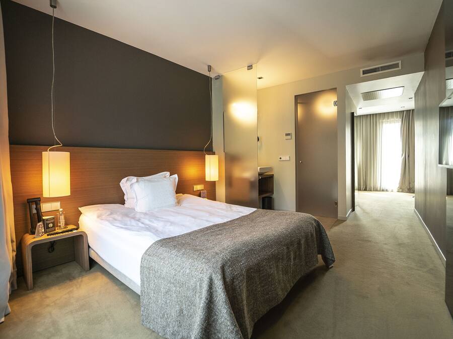 A hotel room with a large bed, bedside lamps, a nightstand, a TV, and a hallway leading to a bathroom.