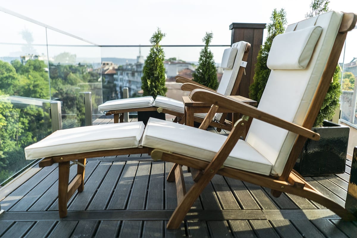 A wooden deck with two lounge chairs and a small table, overlooking a cityscape with trees and buildings.
