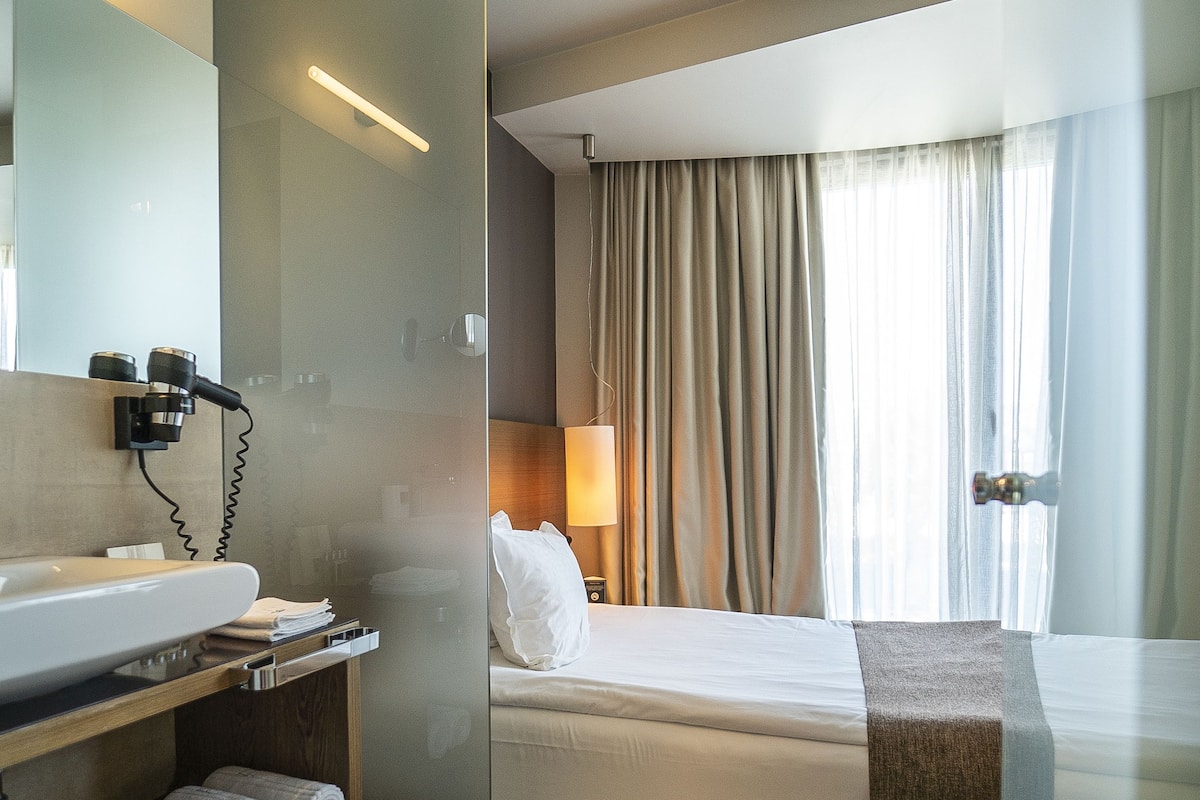 A hotel room with a glass partition, a bed, a bedside table with a lamp, a bathroom with a sink and a hairdryer, and a window with curtains.
