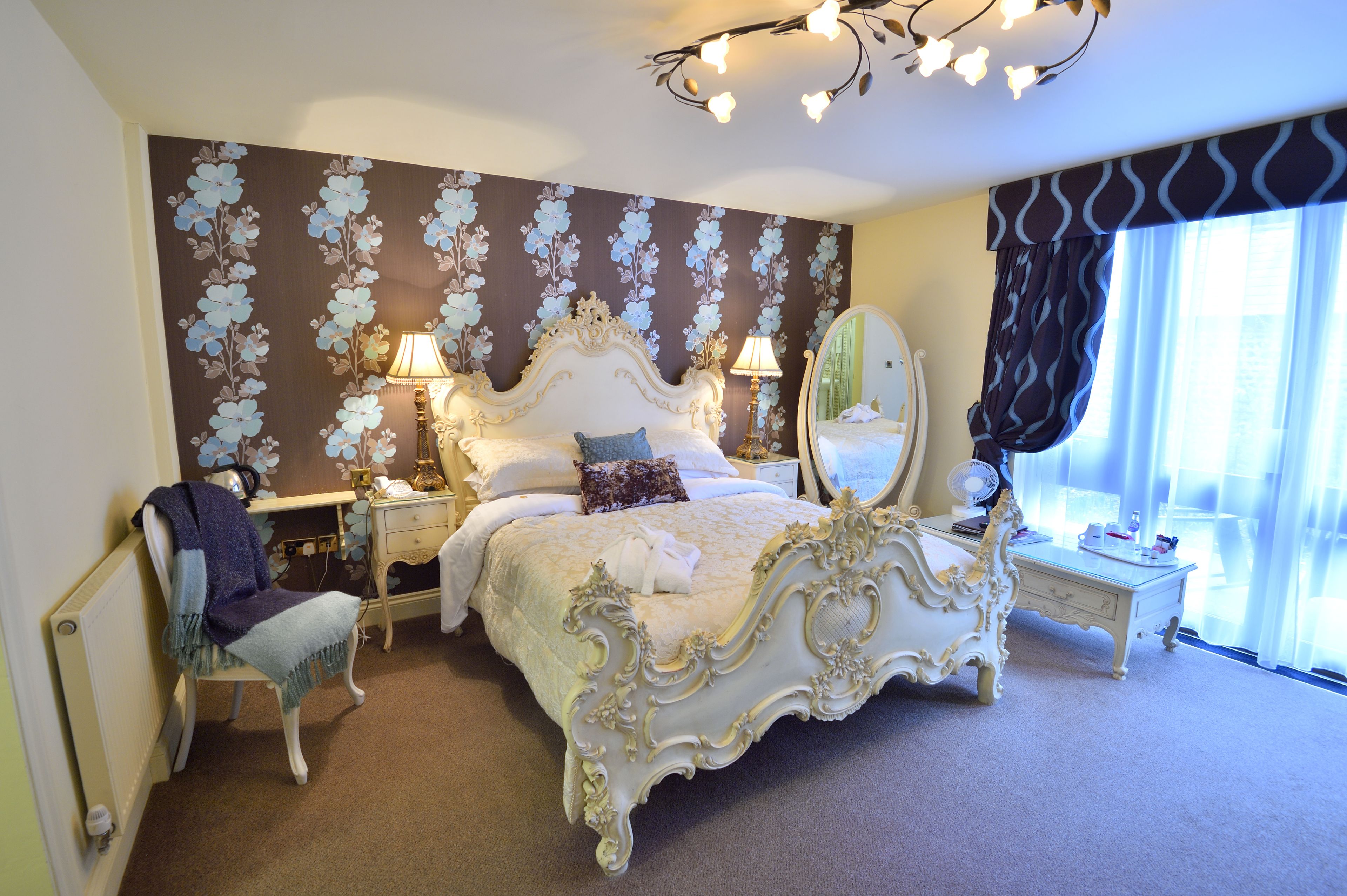 A bedroom with a large ornate bed, a bedside table with a lamp, a chair with a blanket, and a mirror on the wall.