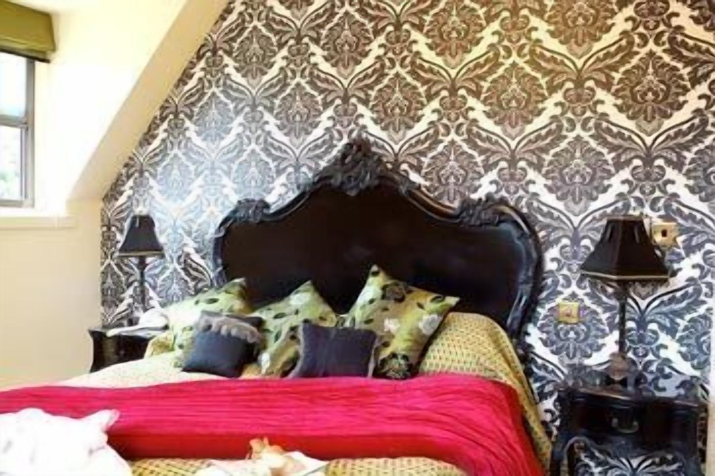 Un dormitorio con papel tapiz estampado, cabecero oscuro y colcha roja.