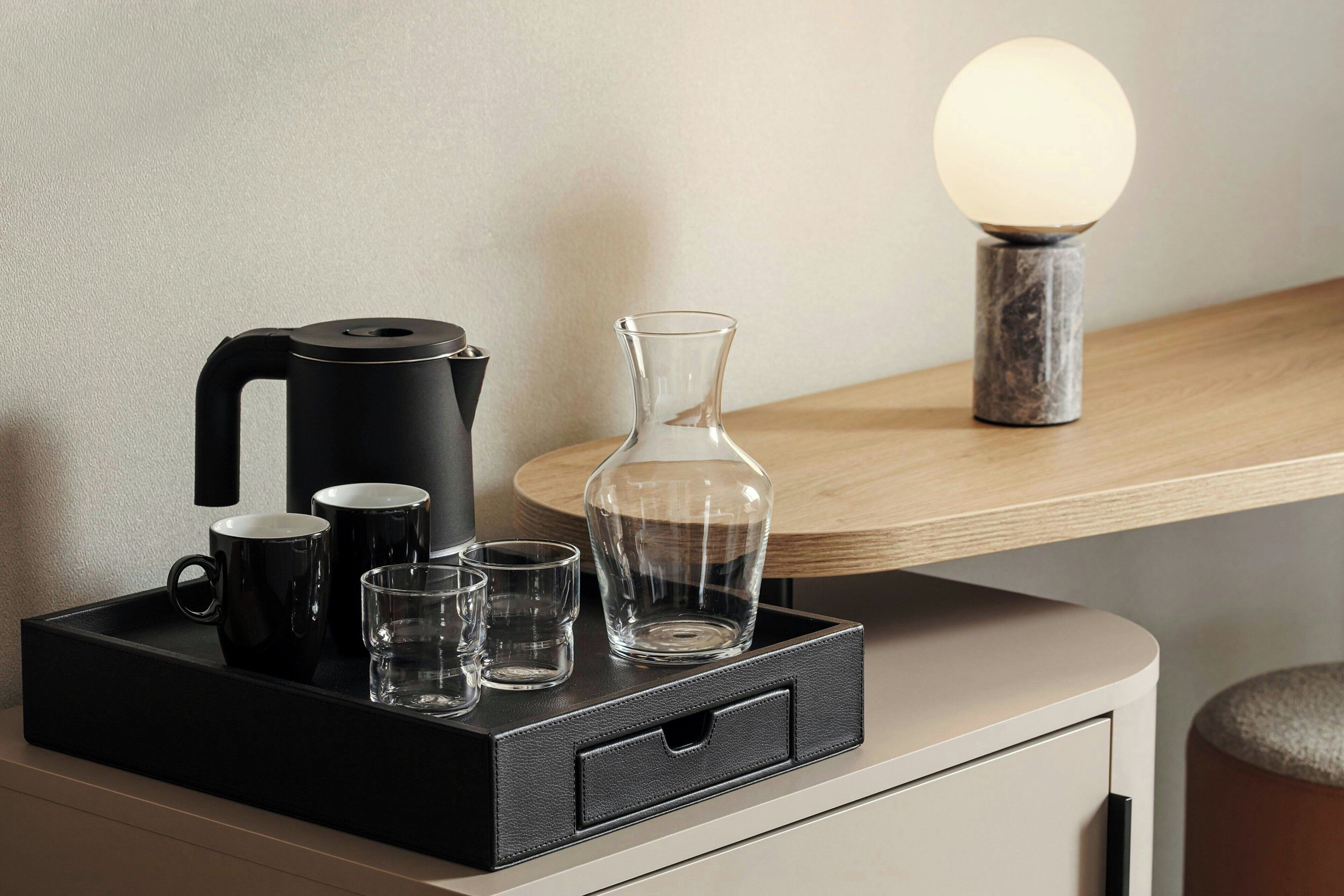 Una bandeja negra con una cafetera, dos tazas negras y tres vasos transparentes sobre una mesa de madera.