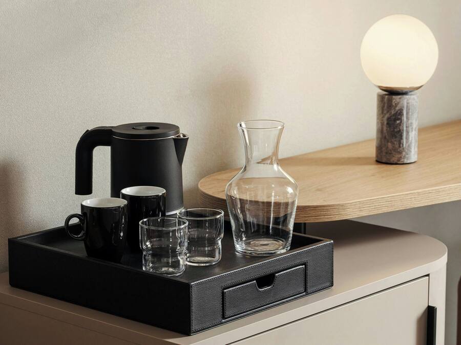 Una bandeja negra con una cafetera, dos tazas negras y tres vasos transparentes sobre una mesa de madera.