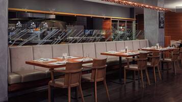 A modern restaurant with wooden tables and chairs, a seating area with cushioned benches, and a view of the kitchen and stairs in the background.