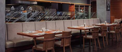 Un restaurante moderno con mesas y sillas de madera, una zona de estar con bancos acolchados, y vistas a la cocina y las escaleras al fondo.