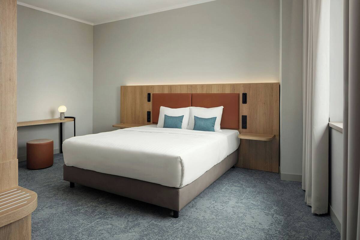 Una habitación de hotel moderna con una cama grande, cabecera de madera, una mesita de noche con lámpara y un pequeño taburete.