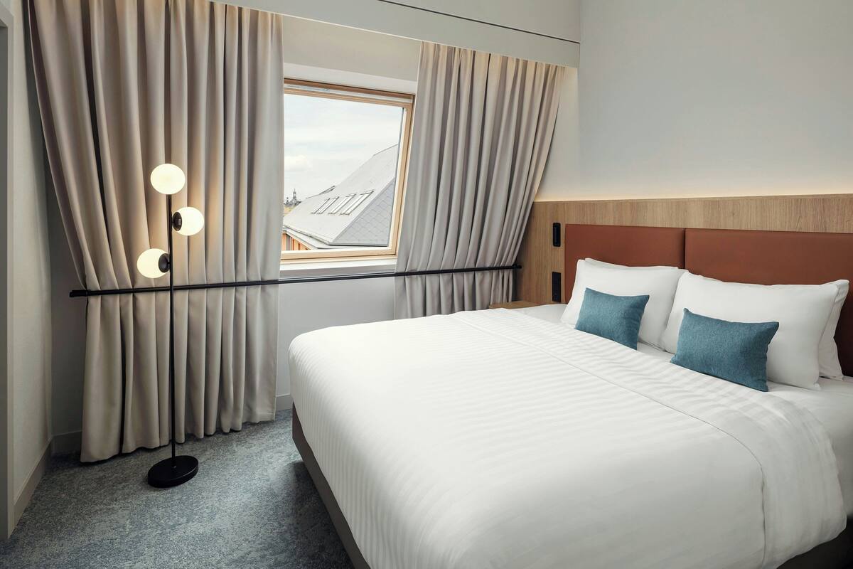 Habitación de hotel con una cama grande, cortinas color beige, una lámpara de pie y vistas a edificios a través de una ventana.