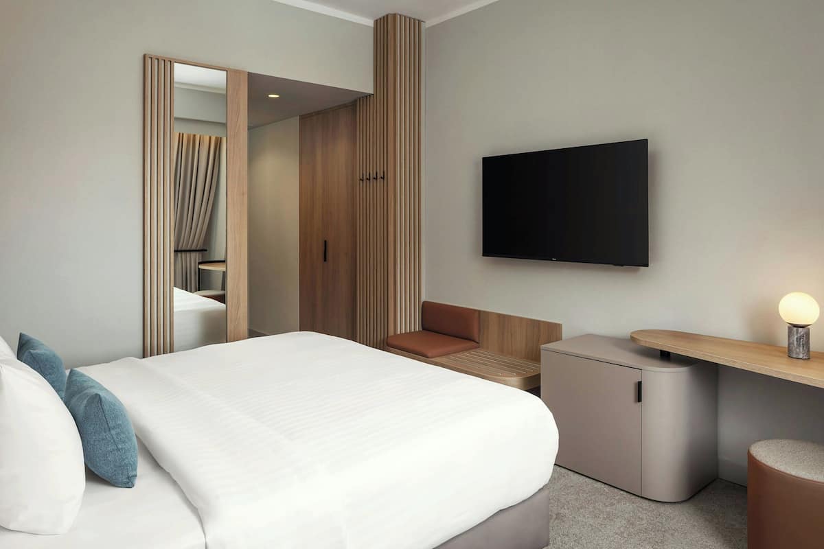 Una habitación de hotel moderna con una cama grande, un televisor montado en la pared, una pequeña zona de estar y un mueble de almacenamiento compacto.