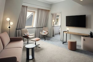 Una habitación de hotel moderna con sofá, dos sillones, una mesita, un otoman redonda, un televisor y un escritorio con lámpara.