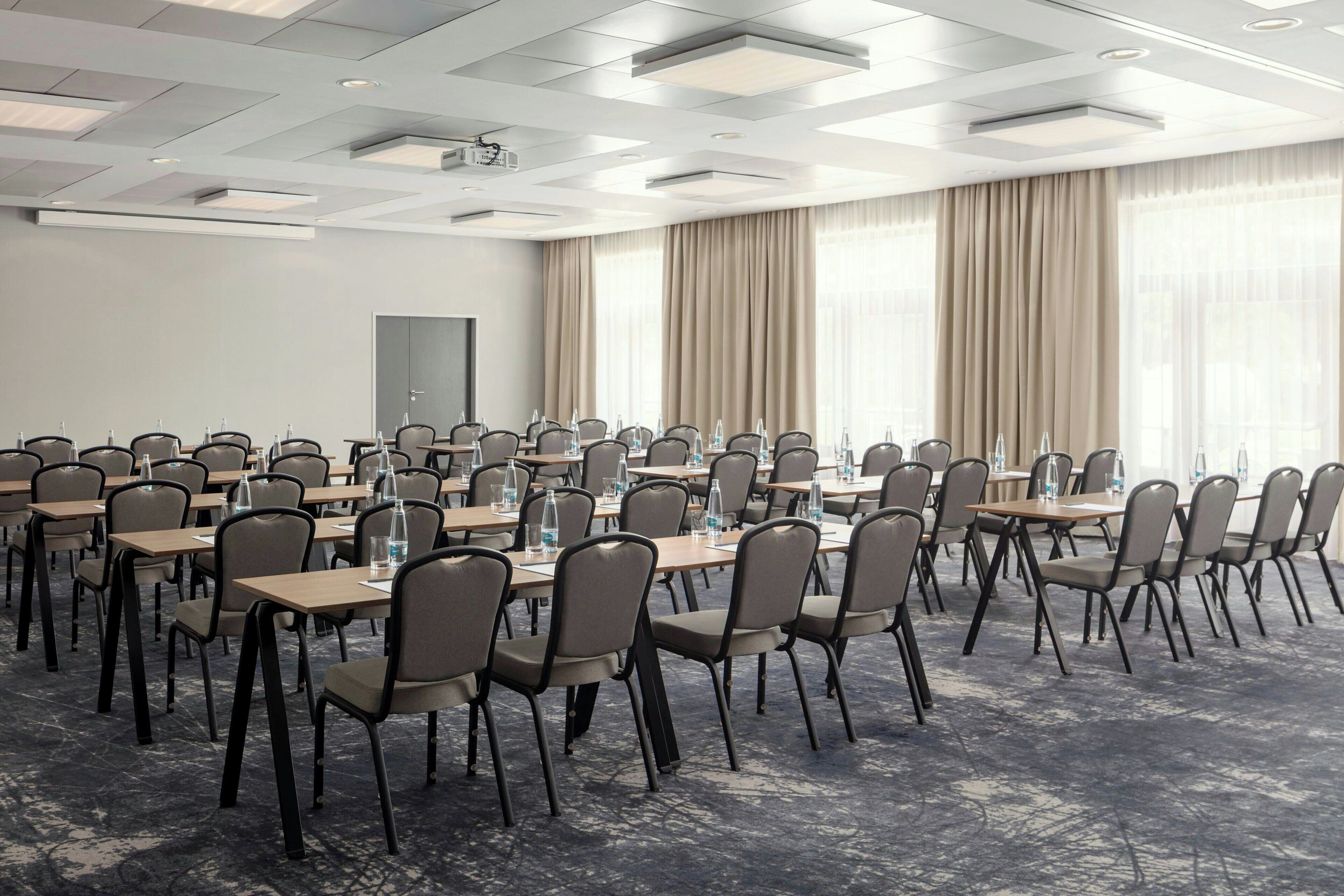 A conference room with multiple tables and chairs, a projector, and large windows with curtains.