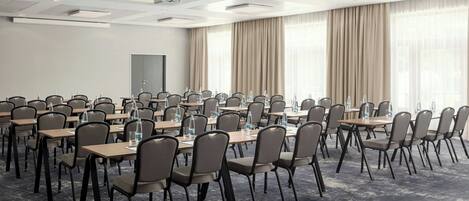 A conference room with multiple tables and chairs, a projector, and large windows with curtains.