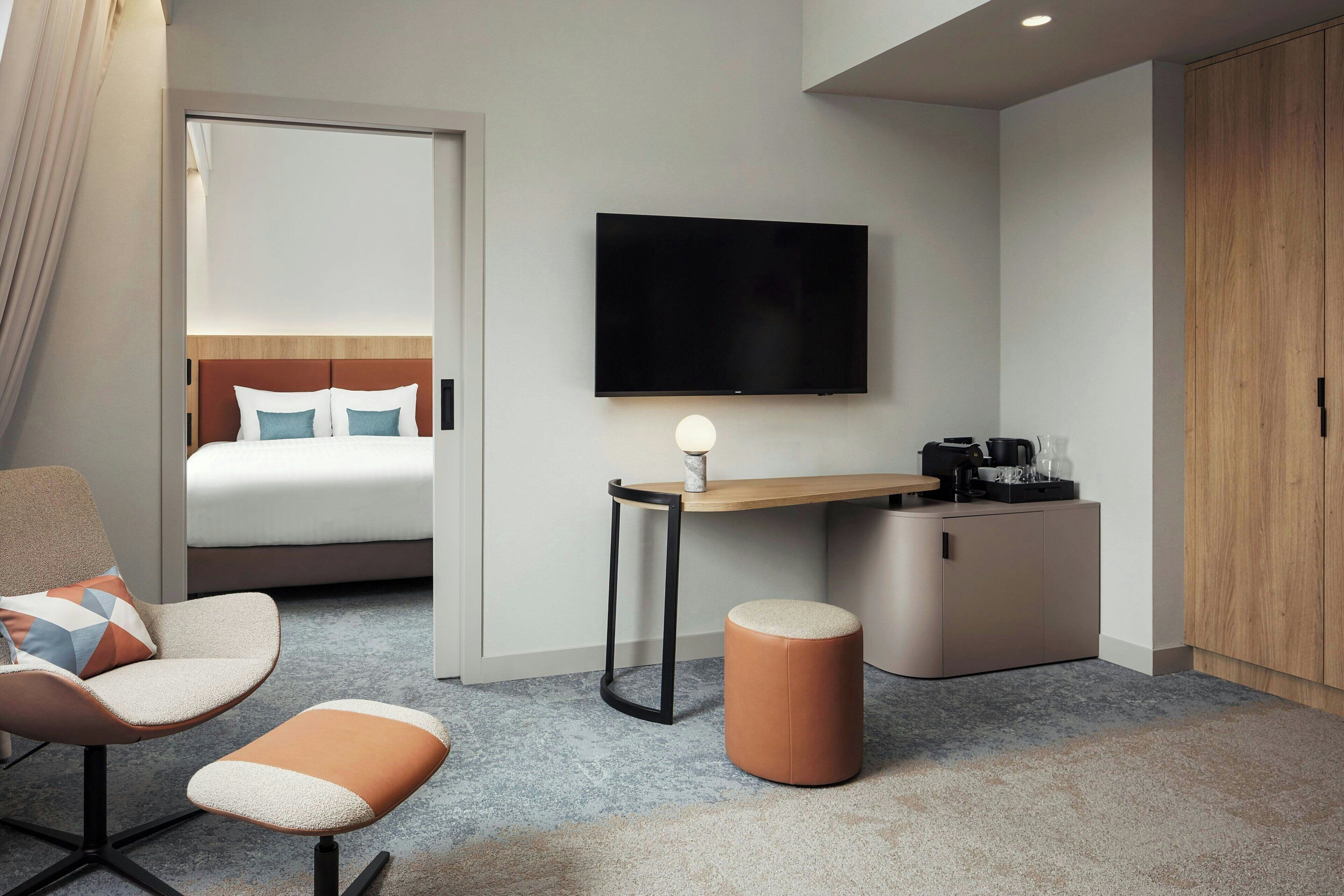 Habitación de hotel con cama, escritorio con televisor, una silla, un pequeño banquito y un armario.