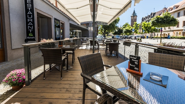 Un espacio exterior para comer con sillas y mesas de mimbre, un letrero para restaurante y bar, y vistas a la calle y edificios al fondo.