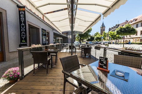 An outdoor dining area with wicker chairs and tables, a sign for a restaurant and bar, and a view of the street and buildings in the background.