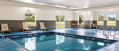 An indoor swimming pool with clear lane markings, wooden lounge chairs, and large windows offering views of the outdoors.