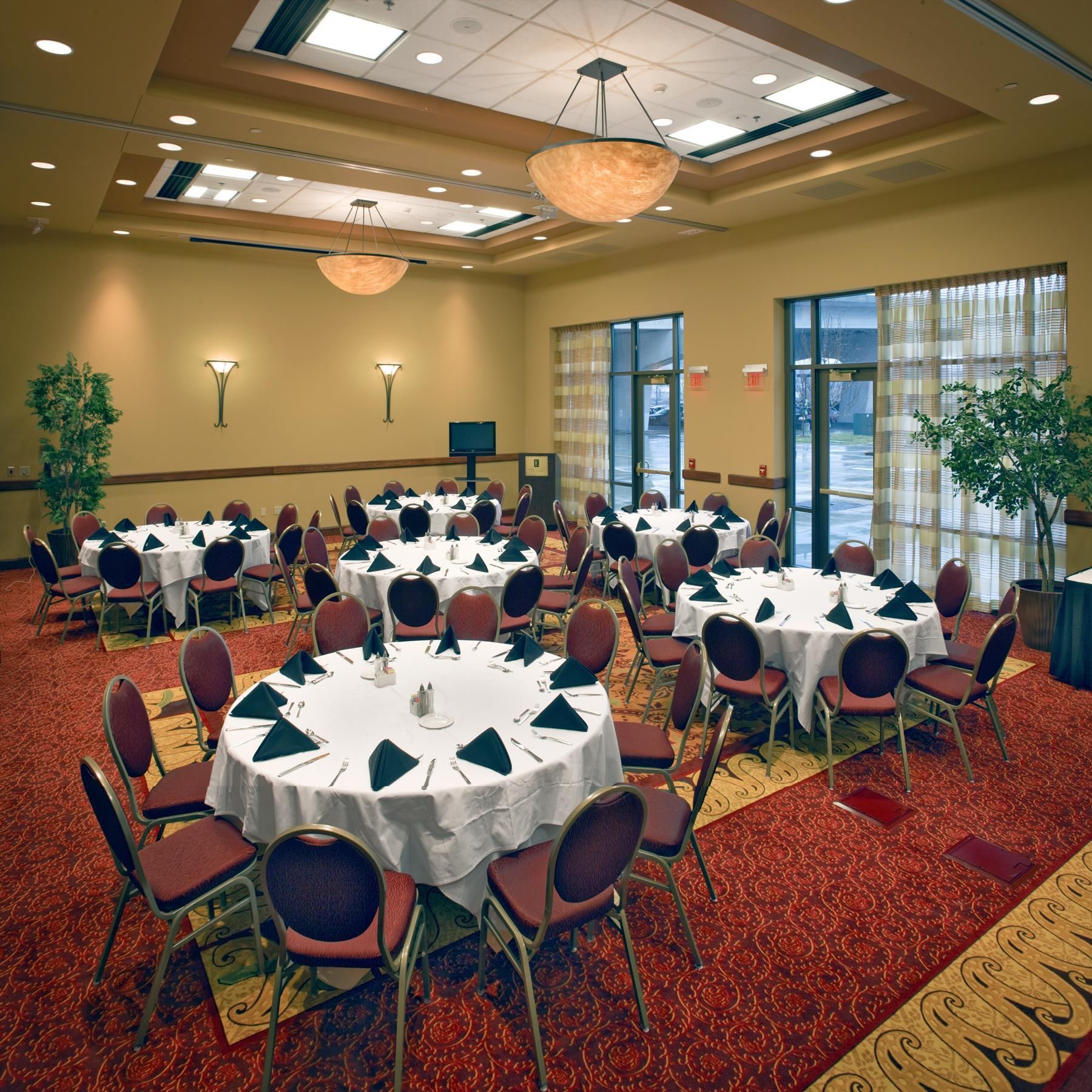 A conference room with multiple tables and chairs, arranged in a circular pattern, with a carpeted floor and a large window providing natural light.