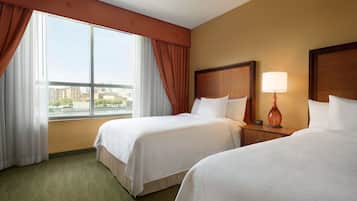 Habitación de hotel con dos camas, una ventana con cortinas, una mesita de noche con lámpara y vistas a edificios.