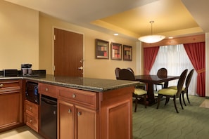 A hotel room with a kitchen area featuring wooden cabinets, a granite countertop, and a built-in microwave. There is a dining table with chairs and a chandelier above.