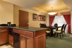 Habitación de hotel con cocina equipada con armarios de madera, encimera de granito y microondas integrado. Se observa una mesa de comedor con sillas y una araña colgante.