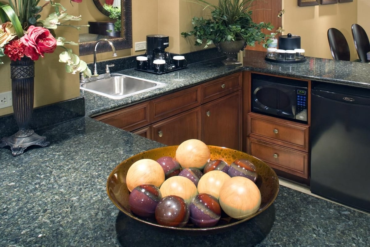Una cocina con encimeras de granito, cafetera, lavamanos y un cuenco de fruta.