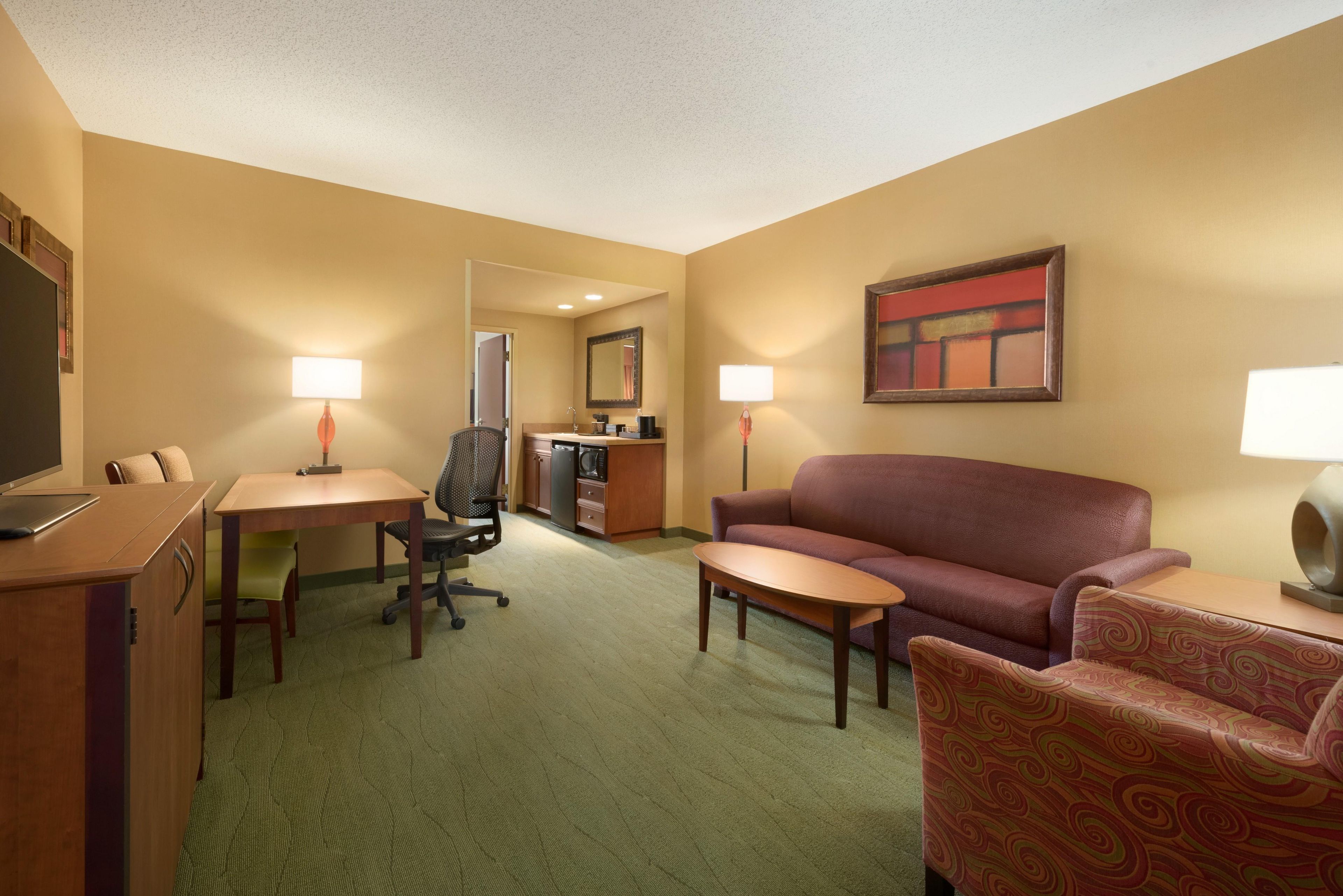 Habitación de hotel con escritorio, silla de oficina, sofá y sillón. Cuenta con una kitchenette con microondas y cafetera. En la pared hay un cuadro, y un televisor está en el lado izquierdo.