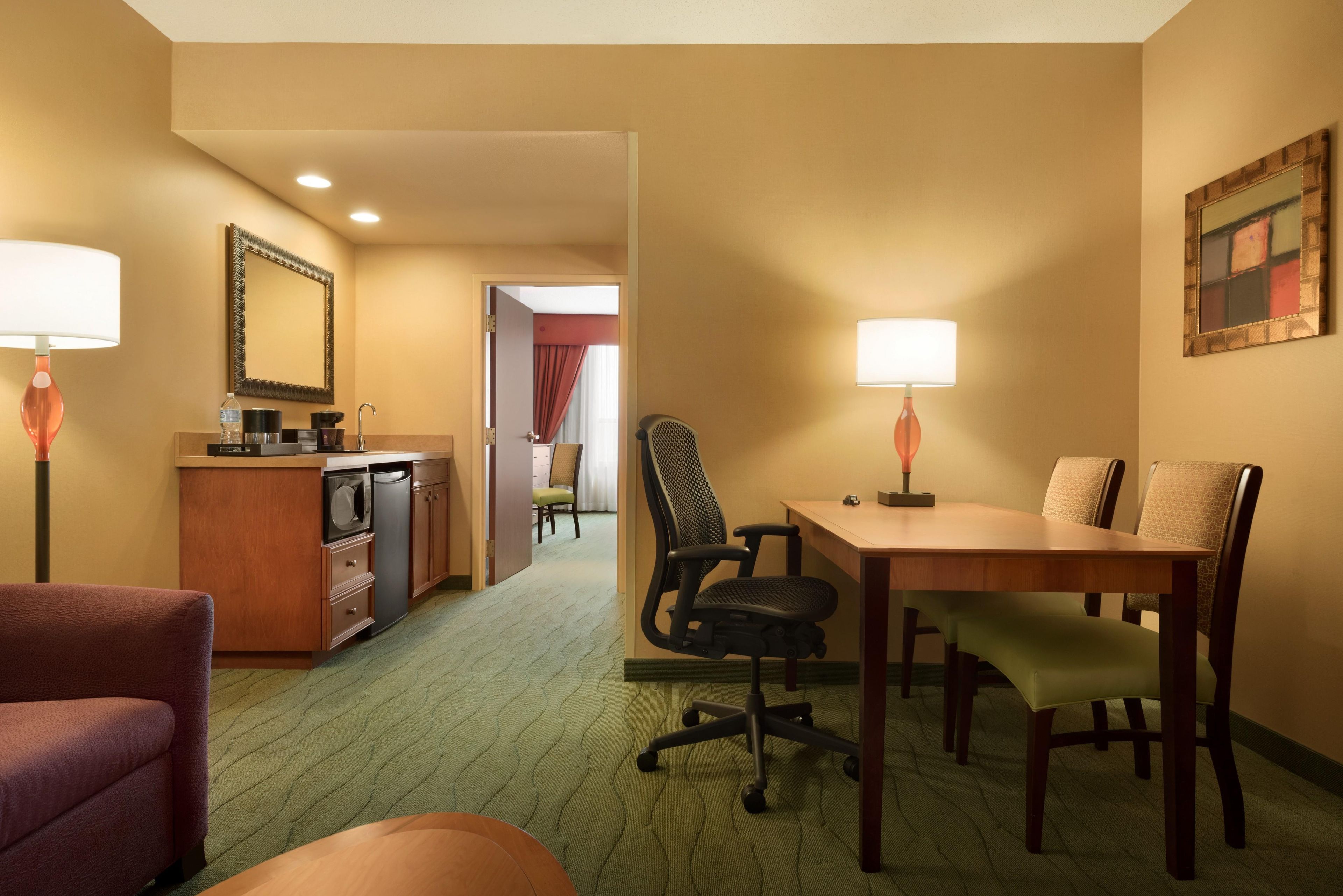 Habitación de hotel con kitchenette, zona de comedor con mesa y sillas, un espejo grande y un cuadro en la pared.