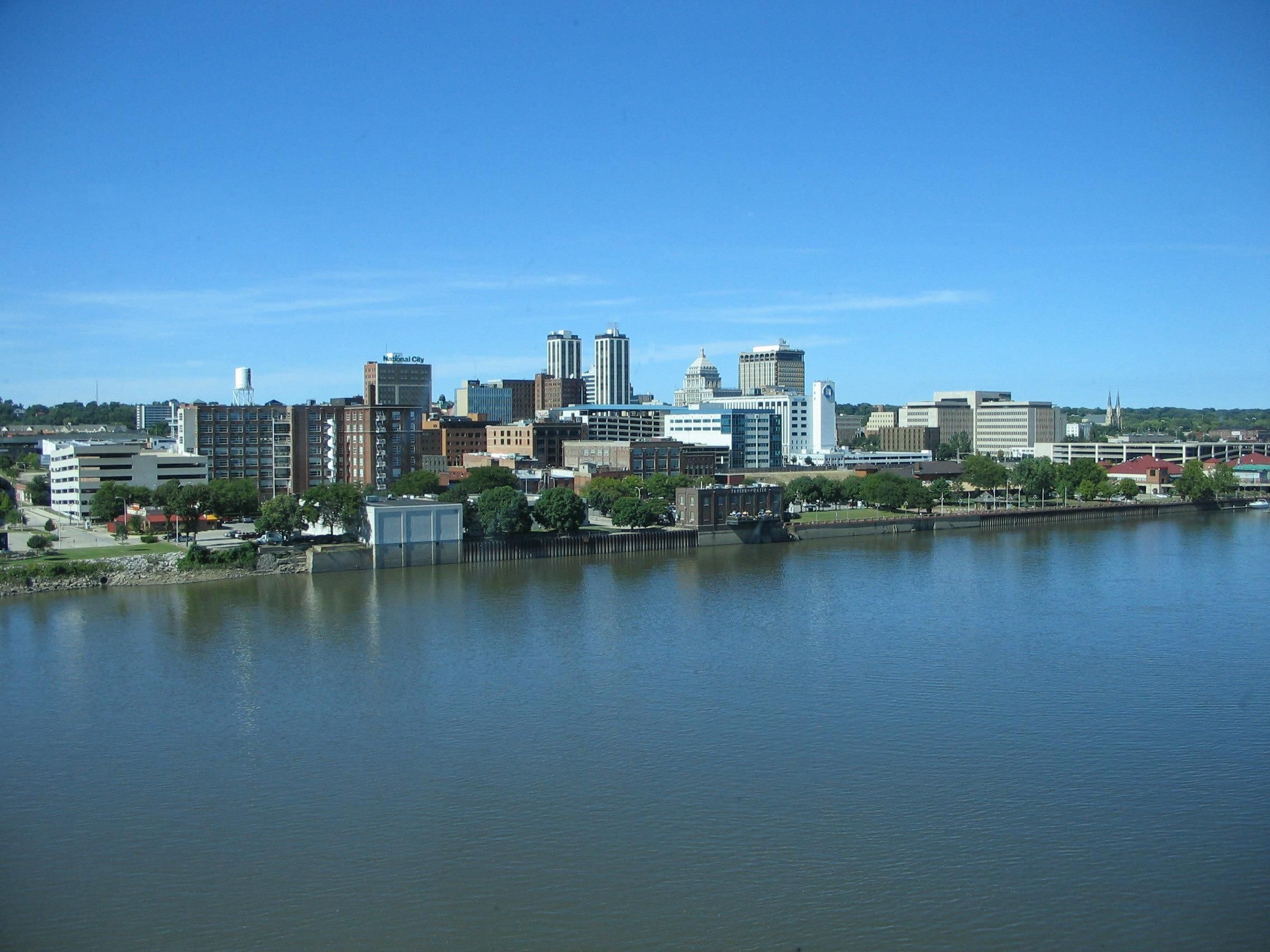 Une vue sur les toits d’une ville, avec différents bâtiments, une rivière au premier plan et un ciel bleu dégagé.