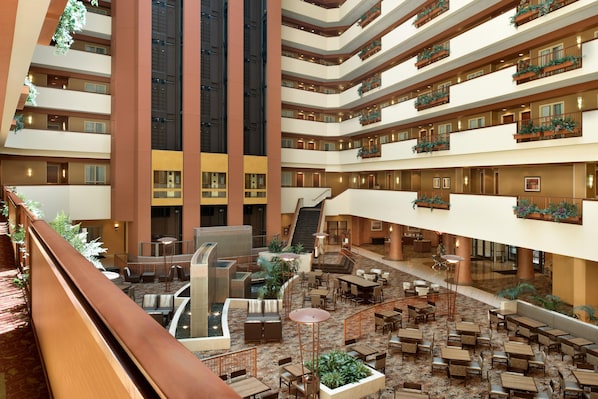 Un vestíbulo de hotel de varios niveles con una escalera central, zonas de estar al aire libre y balcones con plantas.