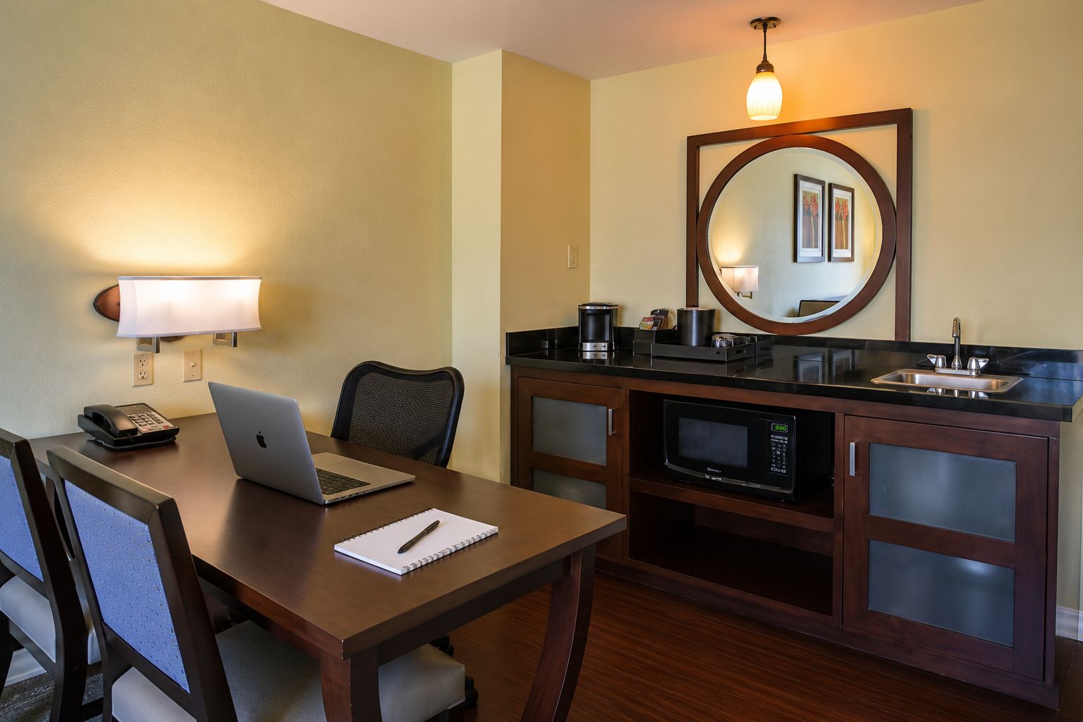 Une chambre d’hôtel équipée d’un bureau en bois, d’un ordinateur portable, d’un téléphone, d’un bloc-notes, d’une chaise, d’un four à micro-ondes, d’une machine à café et d’un lavabo.