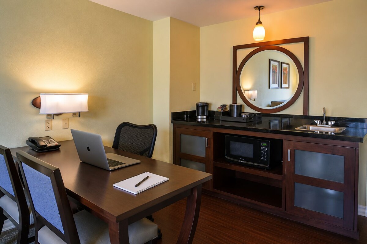 A hotel room with a wooden desk, a laptop, a telephone, a notepad, a chair, a microwave, a coffee maker, and a sink.