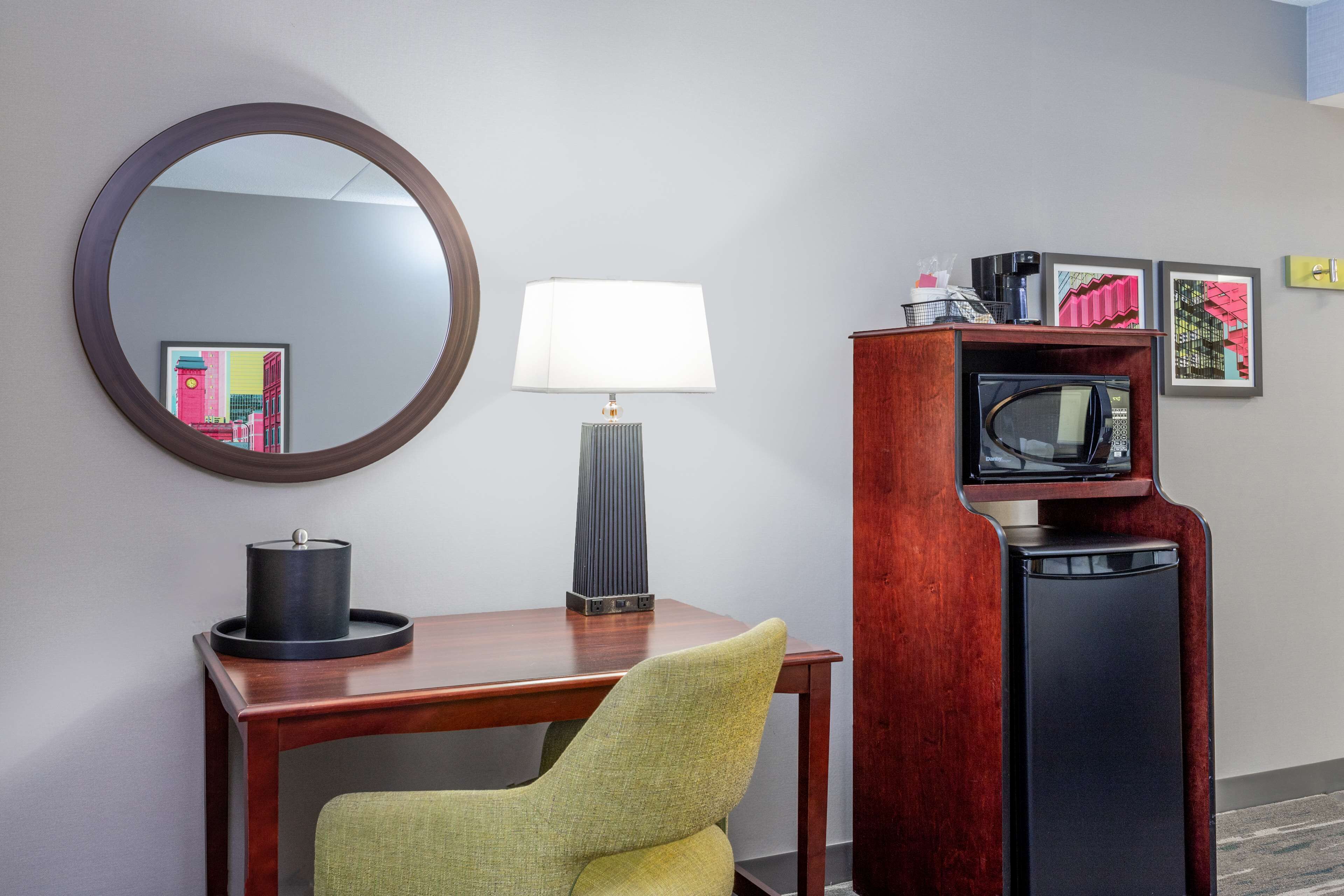 A hotel room with a wooden desk, a green chair, a round mirror, a lamp, a television, and framed artwork on the wall.