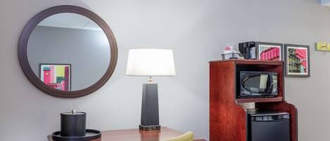 A hotel room with a wooden desk, a green chair, a round mirror, a lamp, a television, and framed artwork on the wall.