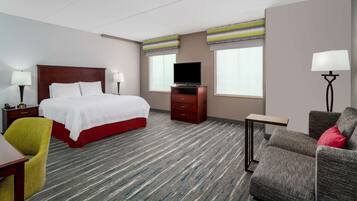 Una habitación de hotel con una cama, un televisor sobre un mueble de madera, un escritorio con lámpara, una silla y un sofá con un cojín rojo.
