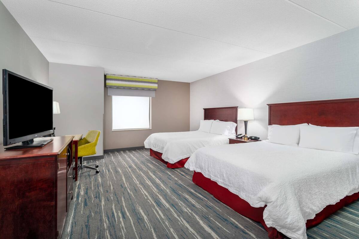 A hotel room with two beds, a desk with a TV, a chair, and a window with blinds.