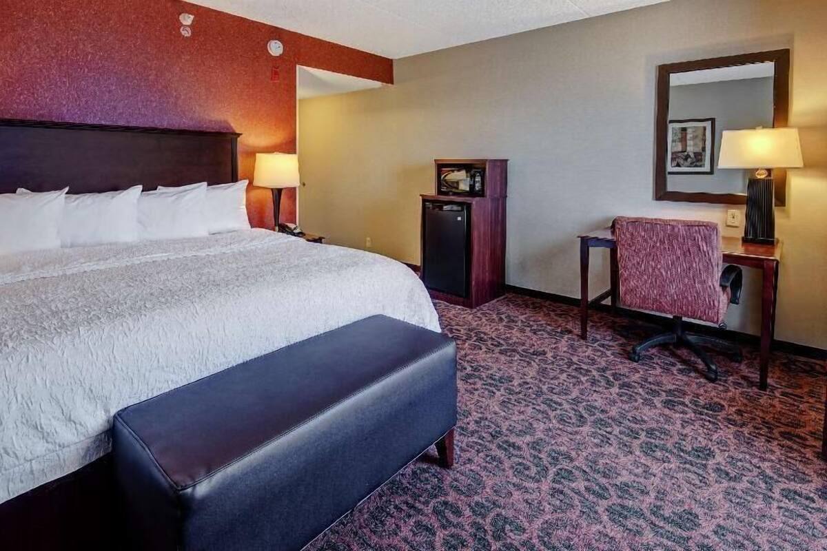 A hotel room with a large bed, a desk, a chair, a small cabinet, and a mirror on the wall.
