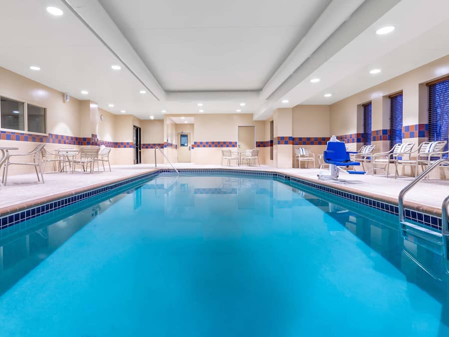 Piscina com bordas em azulejo, rodeada por cadeiras e mesas, e teto com iluminação embutida.