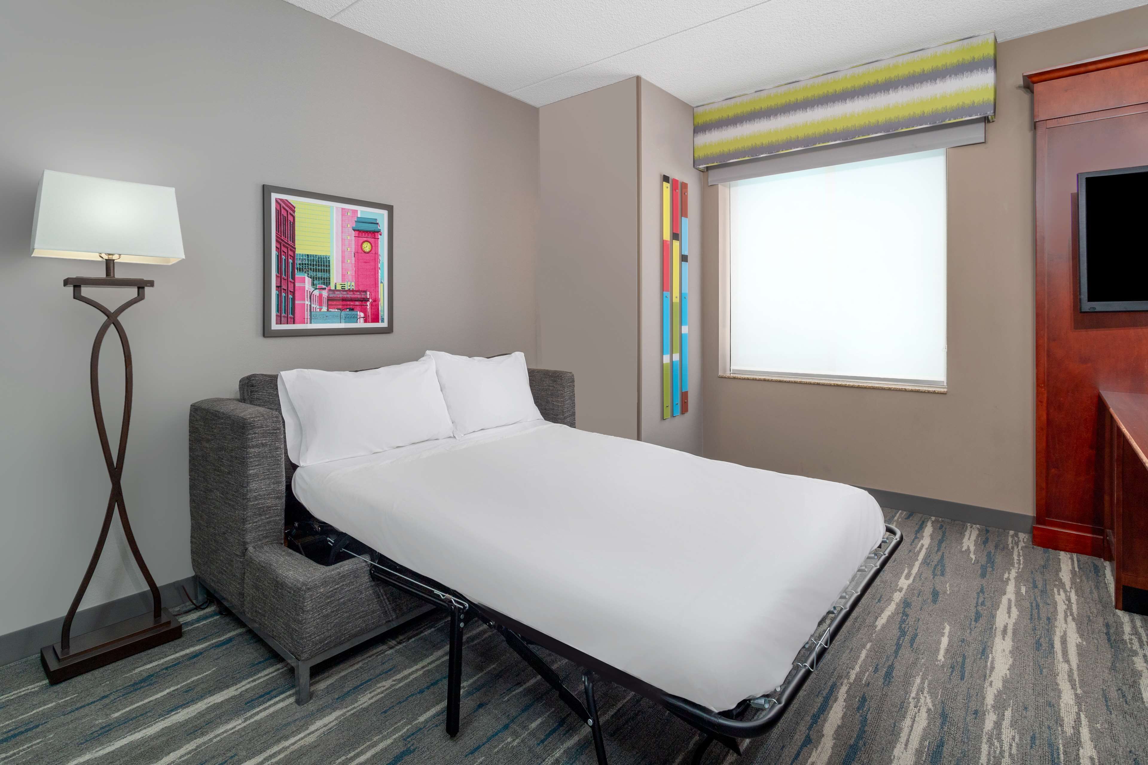Una habitación de hotel con una cama individual, una lámpara de pie, un cuadro abstracto de colores vivos, una ventana con persianas y un televisor sobre un mueble de madera.