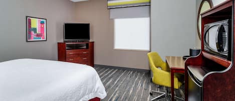A hotel room with a bed, a television, a desk with a chair, a microwave, and a window with blinds.