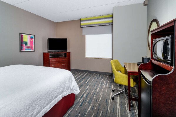 A hotel room with a bed, a television, a desk with a chair, a microwave, and a window with blinds.