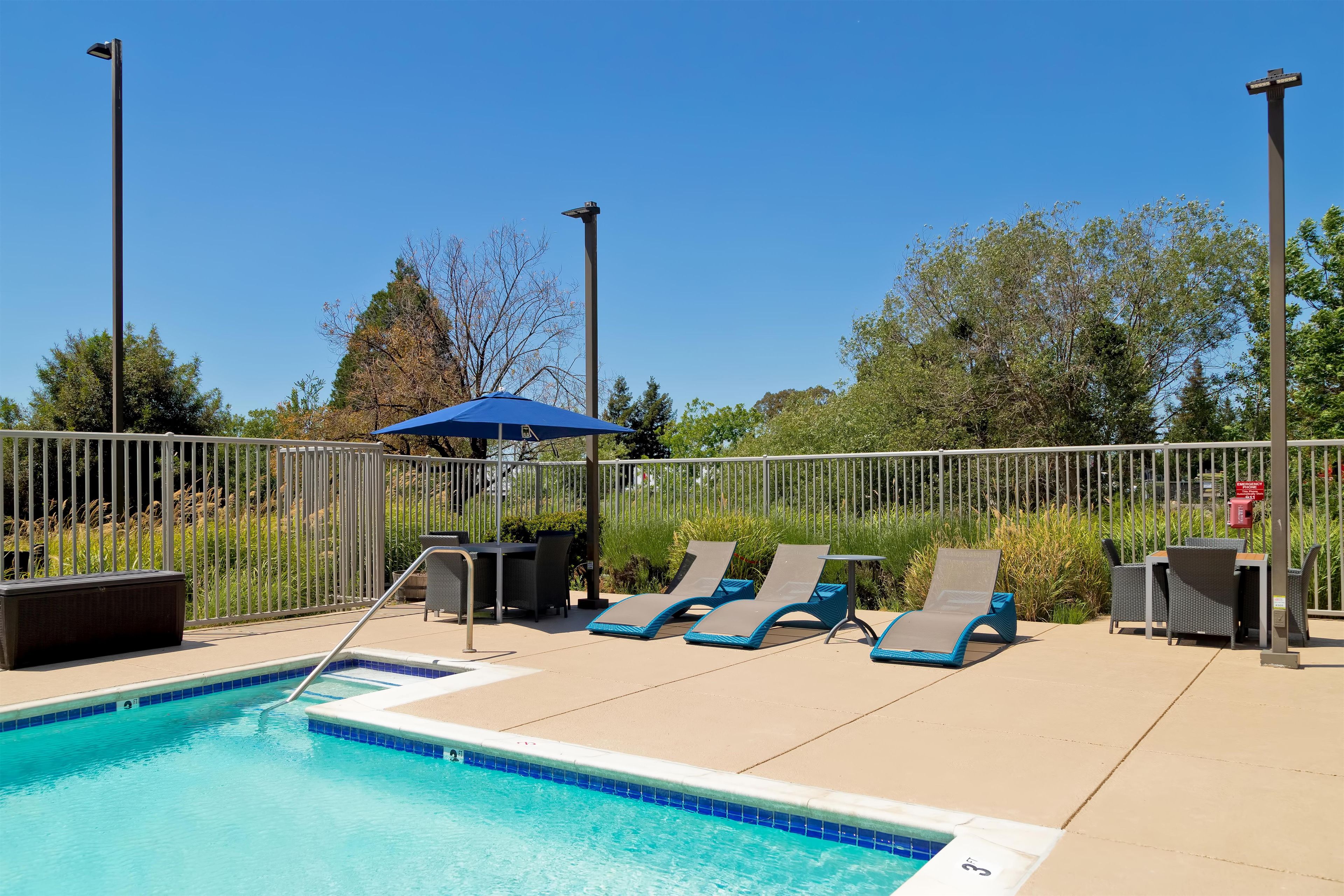 Ein Poolbereich mit Liegestühlen, einem blauen Sonnenschirm und einem eingezäunten Garten.