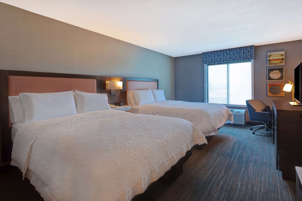 A hotel room with two beds, a desk, a chair, and a large window.